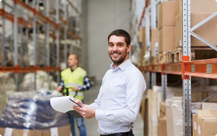 Logistics manager standing in a warehouse aisle holding documents, with storage shelves in the background